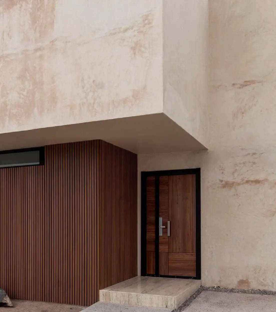 Acceso a Casa Paseo con puerta de madera y marco negro bajo un volumen de aplanado texturizado